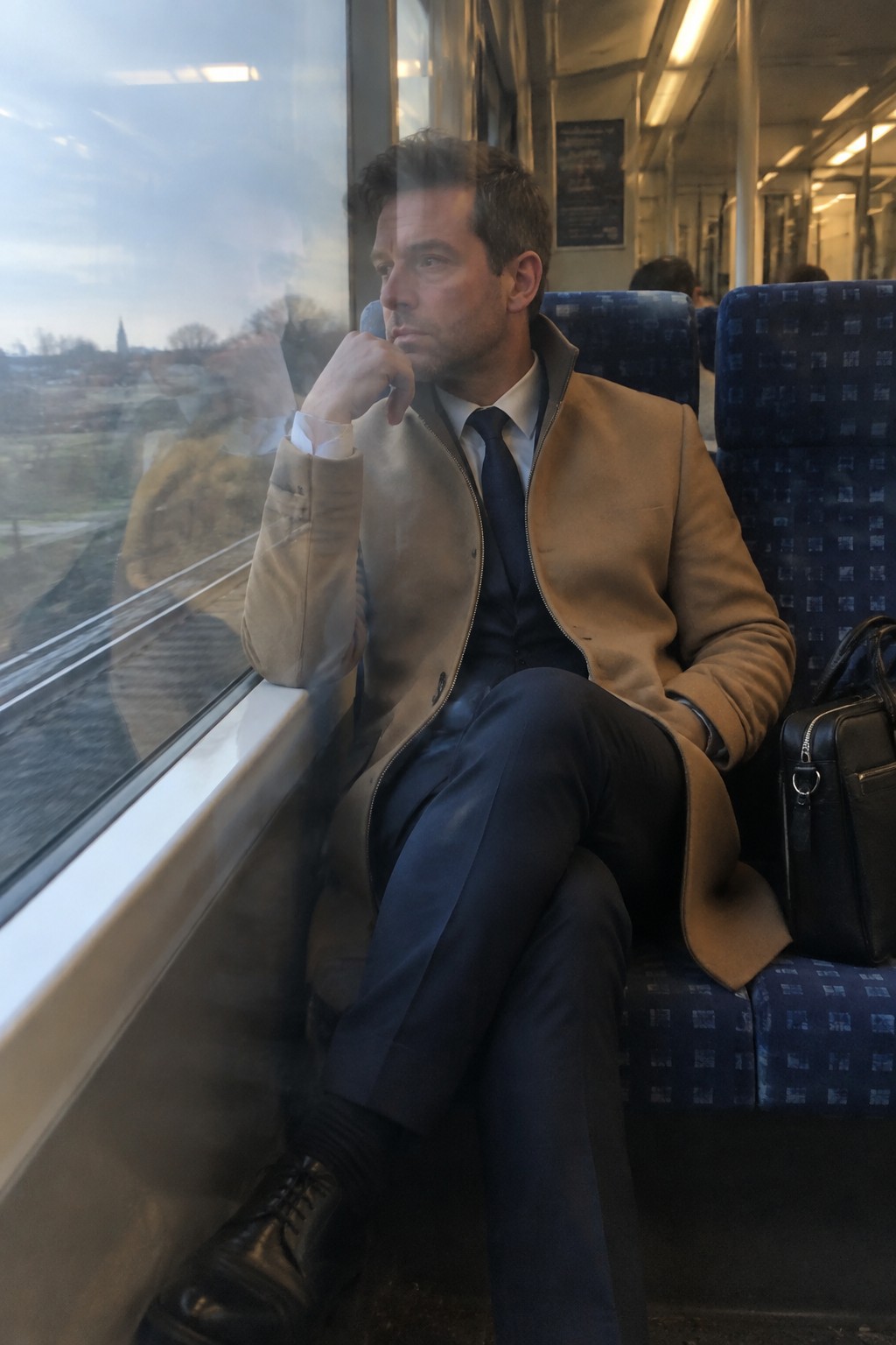 A man wearing a camel suede jacket over a navy suit, white shirt, and navy tie, seated on a train with his legs crossed.