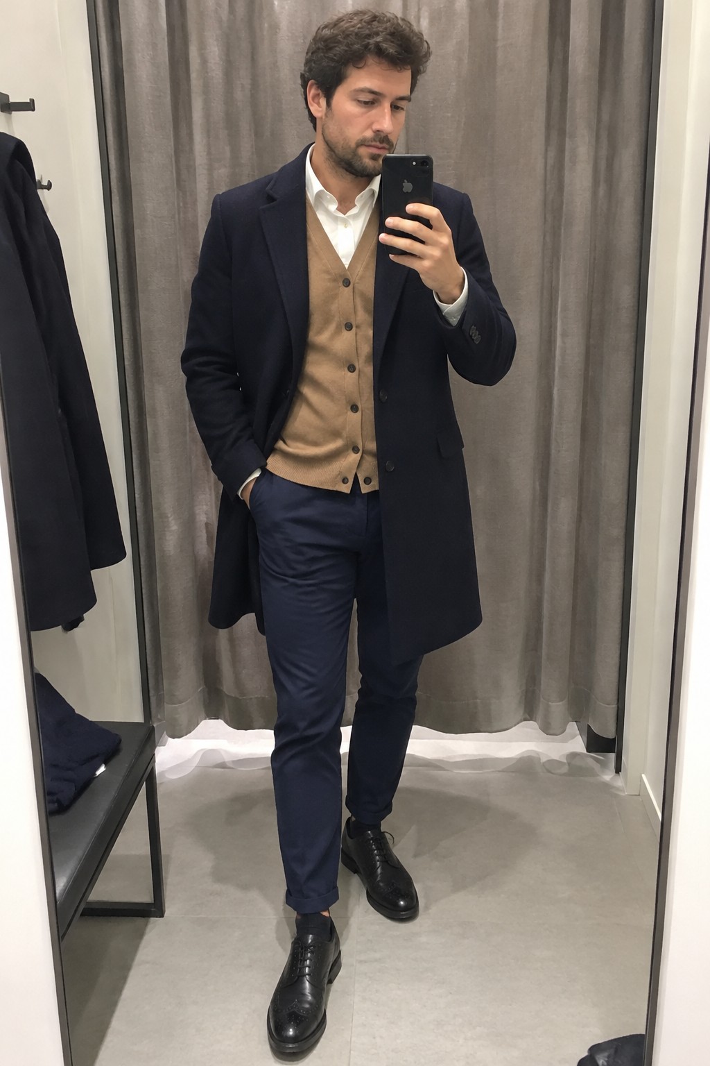 Man wearing a camel buttoned waistcoat over a white shirt with a long navy overcoat, navy trousers, and black brogue shoes.