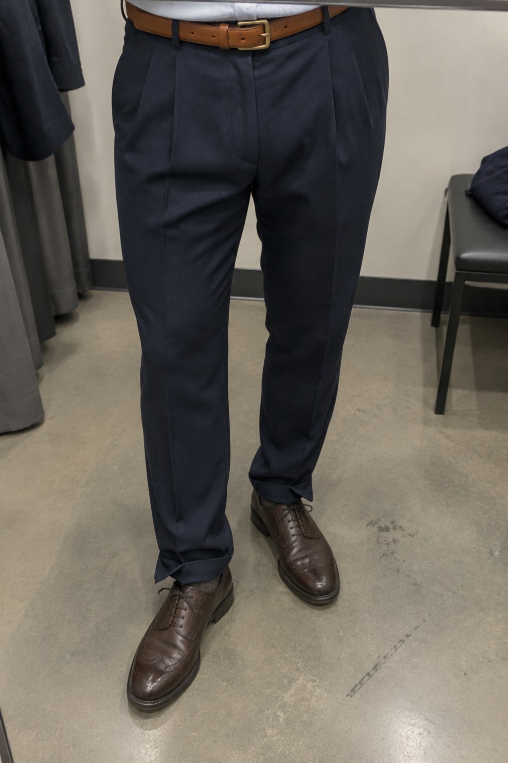 Man wearing navy pleated trousers, a white shirt, camel leather belt, and brown brogue shoes.