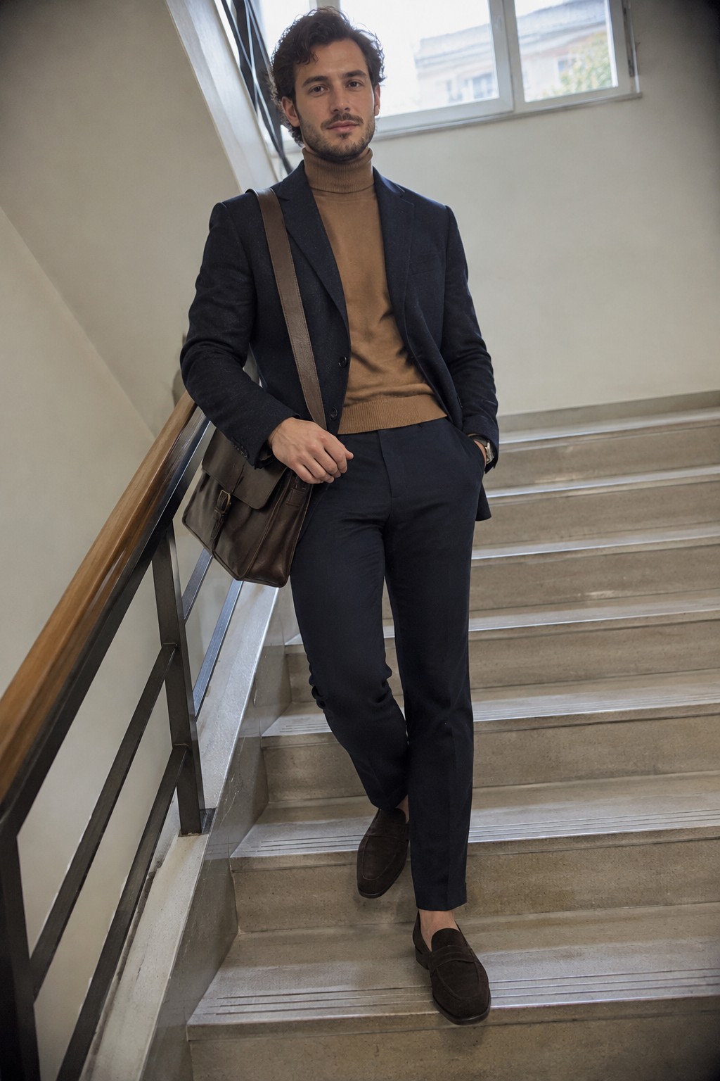A man wearing a navy blazer and trousers, camel turtleneck sweater, dark brown loafers, and a brown leather shoulder bag.